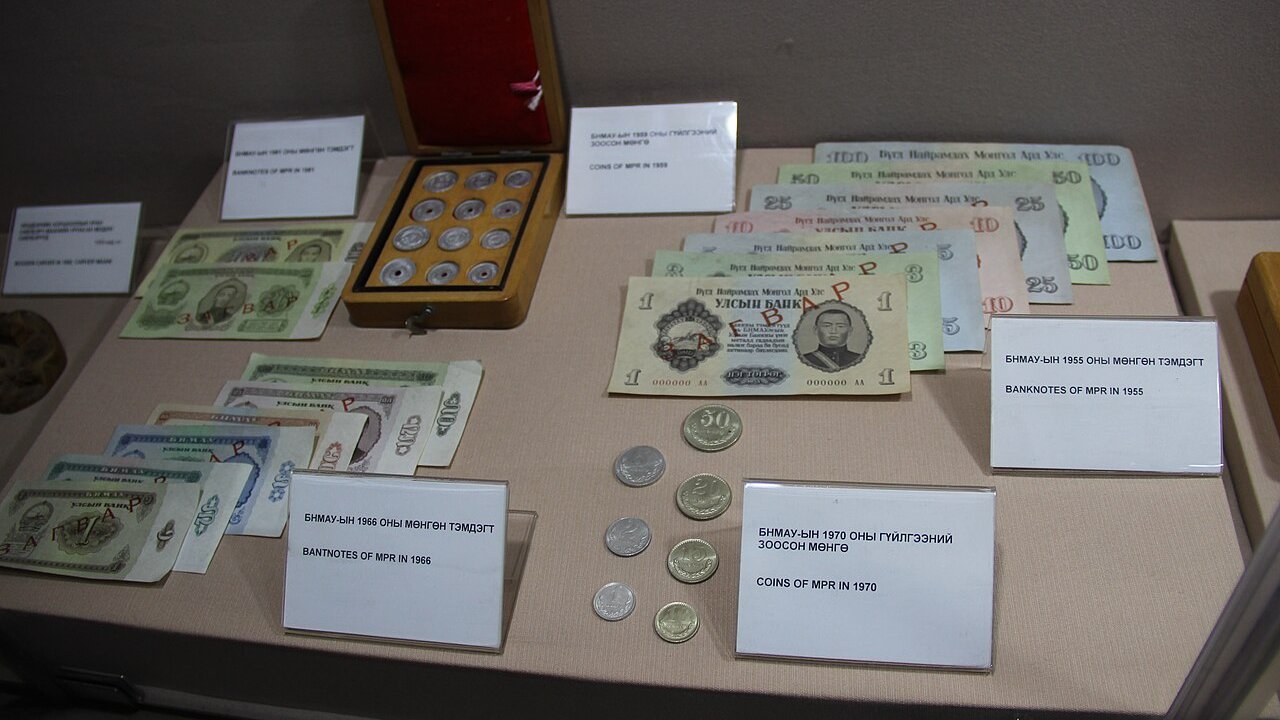Display case of vintage Mongolian banknotes and coins with printed labels describing the currency issued in 1966 and 1970.