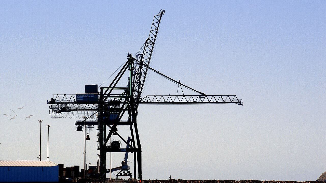 A large dark port container crane silhouetted against a clear sky beside a quay with stacked containers and a blue building.