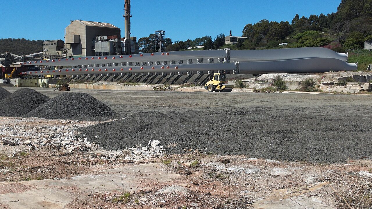 Long grey wind turbine blades lying on a gravel yard beside an industrial plant with a tall chimney.
