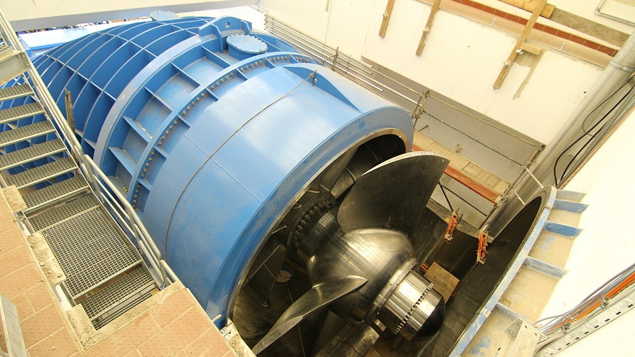 A large blue hydroelectric turbine with a steel runner inside a concrete powerhouse, with access stairs and railings beside it.