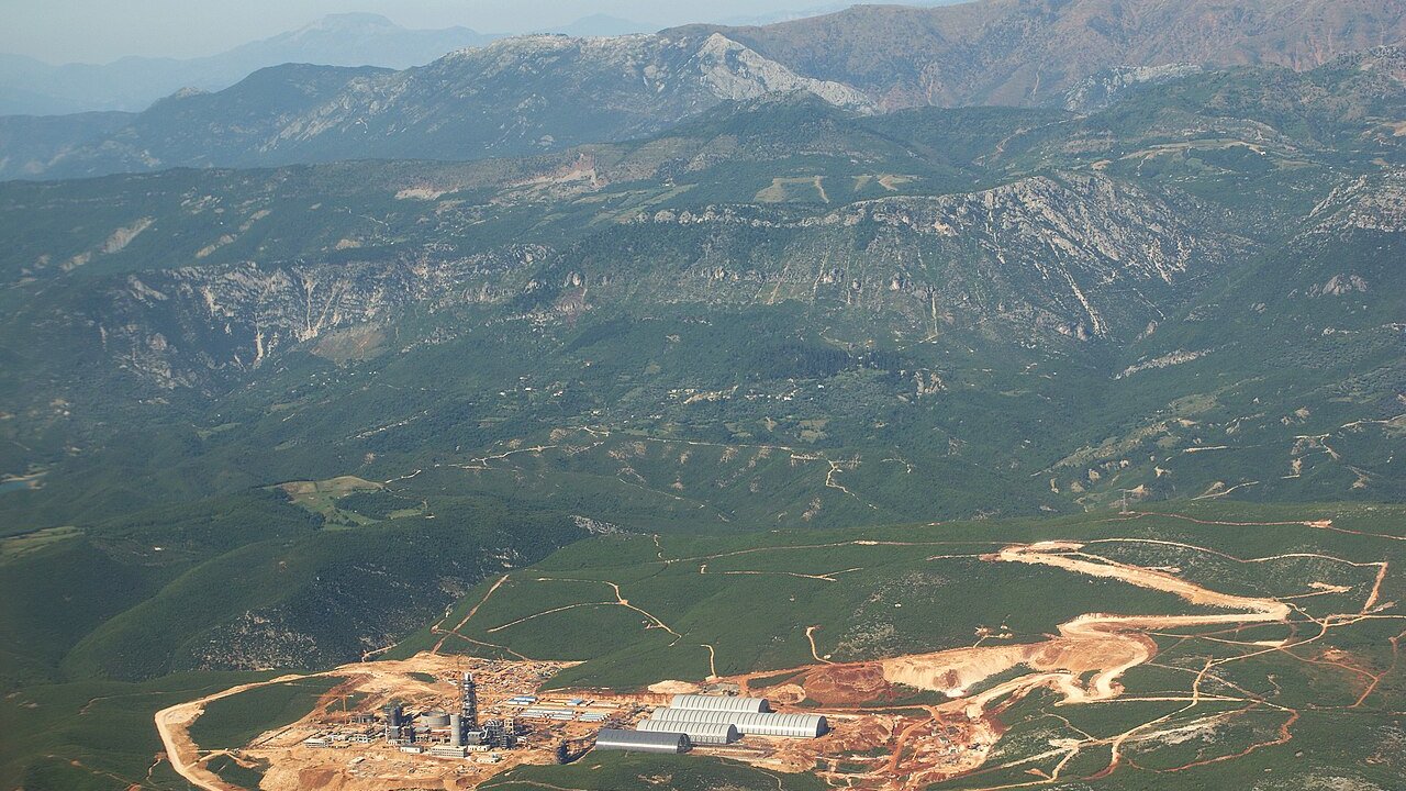 An aerial view of a cement plant with silos and buildings set in green hills, with quarry tracks cut into the slopes.