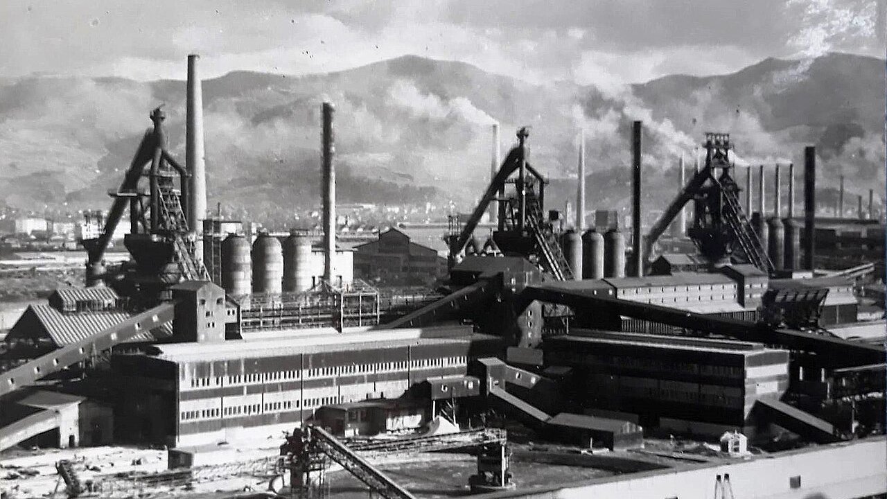 A black-and-white view of an old steelworks with blast furnaces, chimneys, smoke and industrial buildings against a mountain backdrop.