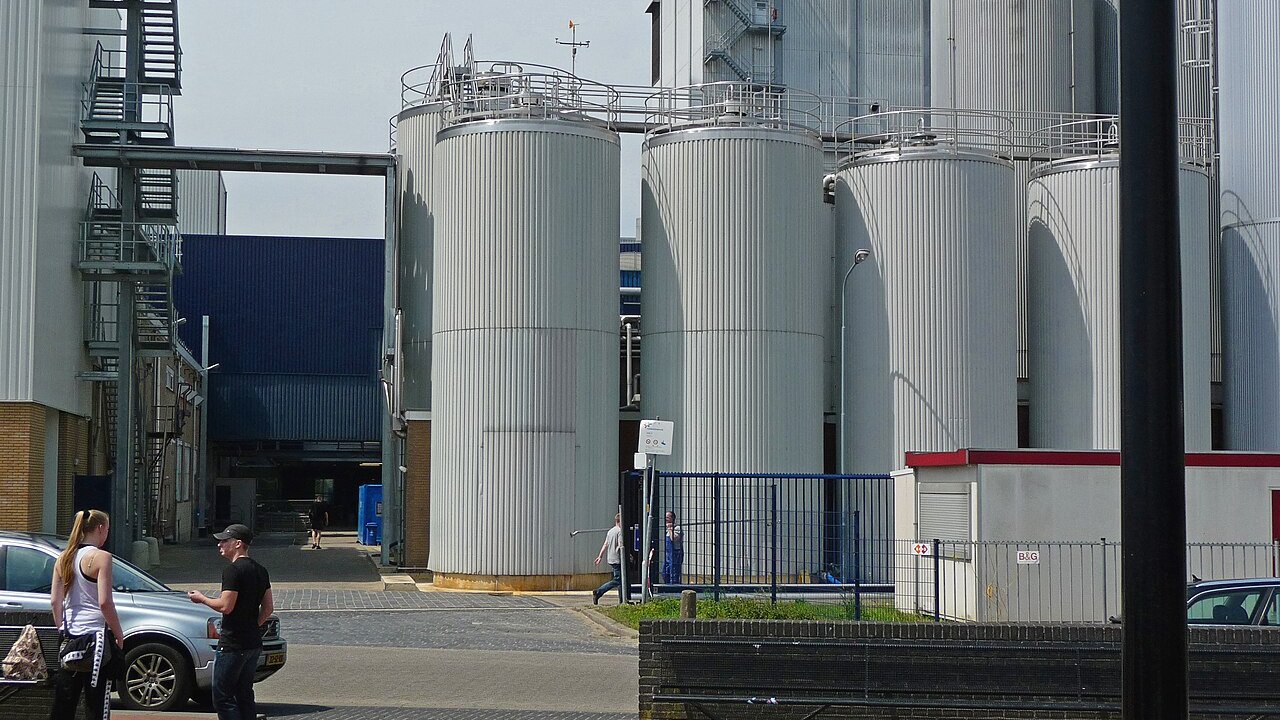 Tall white storage silos of a dairy processing plant beside a paved yard with a blue fence and two people walking.