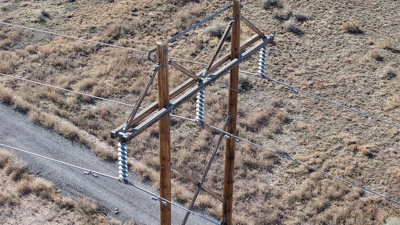 A wooden utility pole carrying overhead power lines with white insulators across dry grassland beside a gravel road.