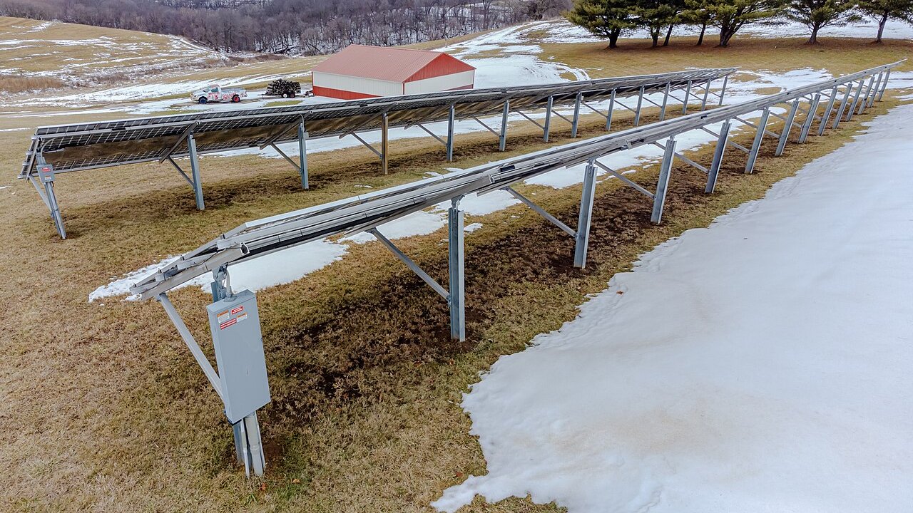 A ground-mounted solar panel array on metal frames across a snowy field, with a red barn in the background