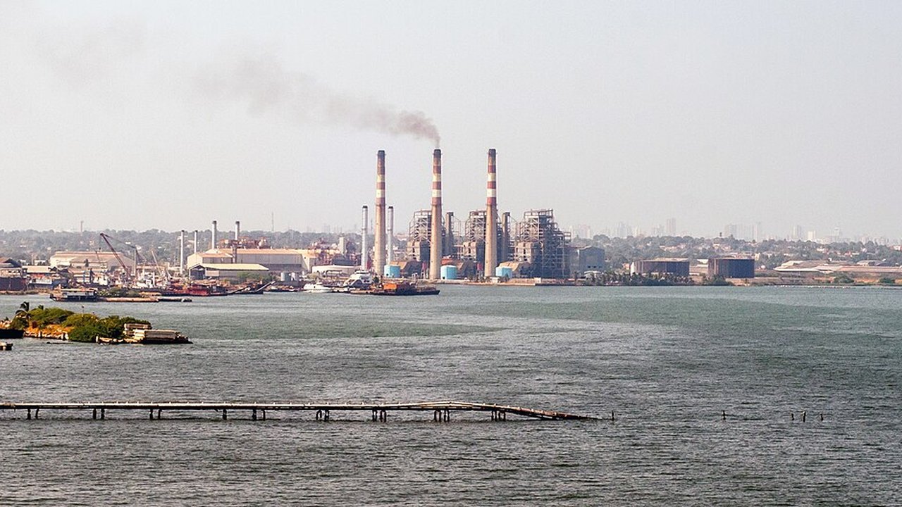 A thermal power plant with tall striped smokestacks emitting smoke beside a harbour and waterfront
