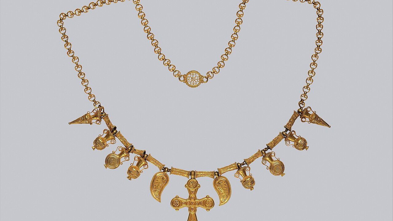 A gold necklace with a central cross pendant, leaf-shaped pieces and filigree ornaments on a plain grey background