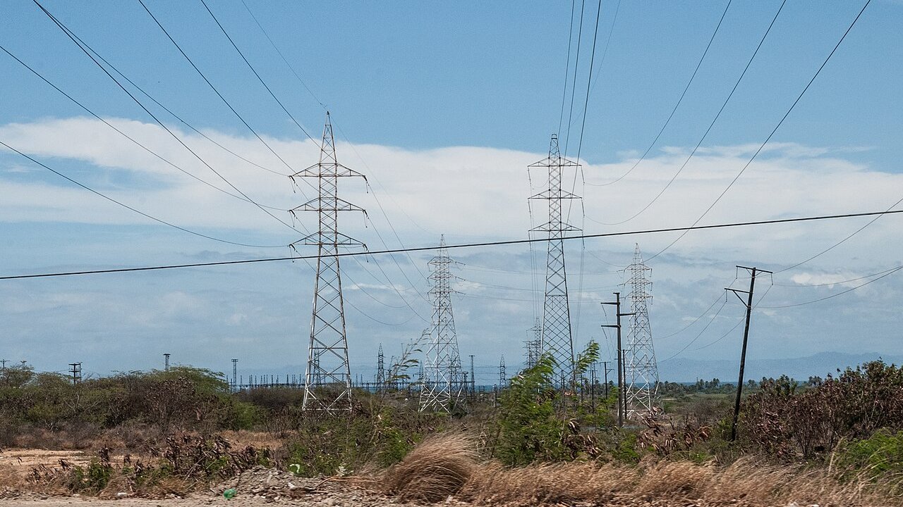 Tall steel electricity transmission pylons and power lines crossing dry scrub land under a blue sky.