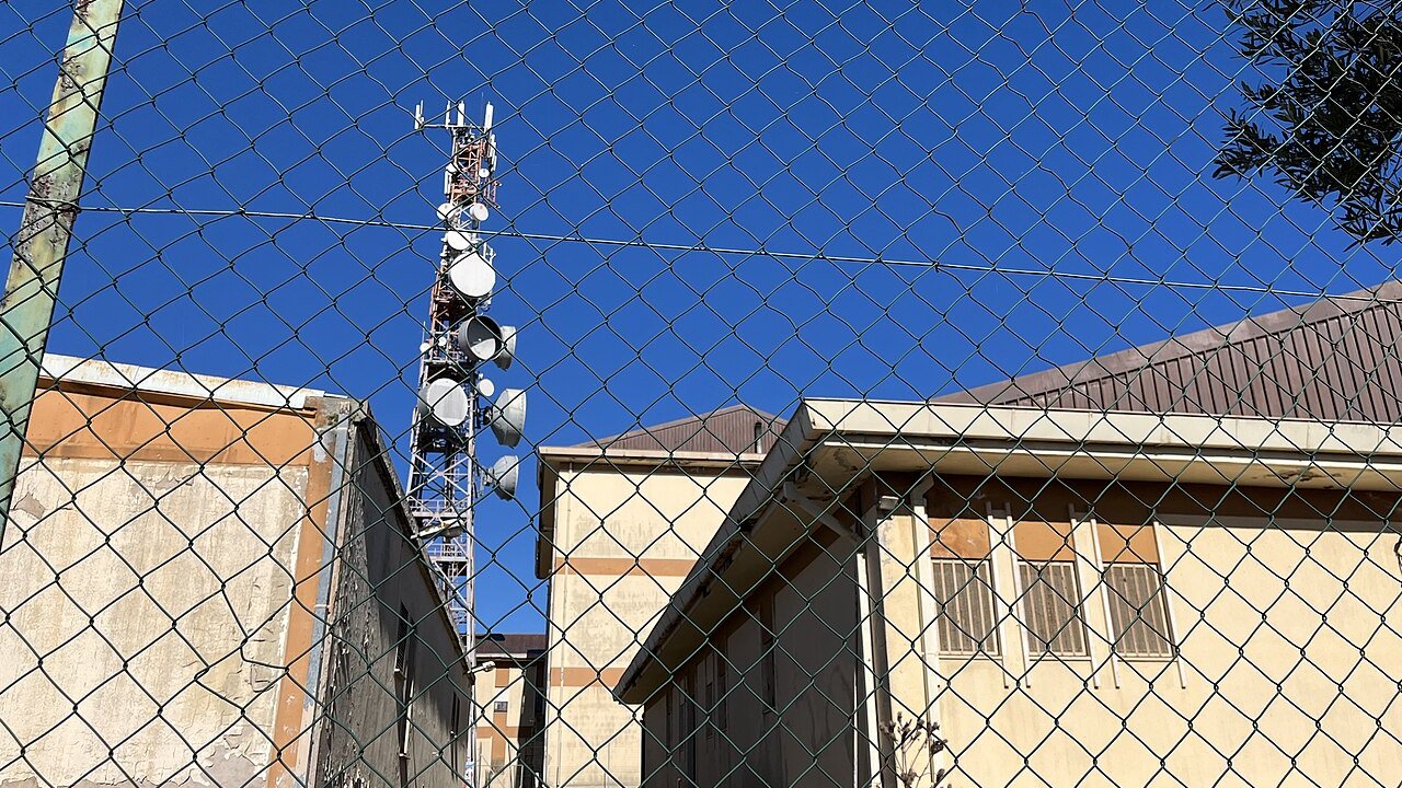 A telecommunications lattice tower with antenna dishes and panels behind a chain-link fence, with buildings in the background.
