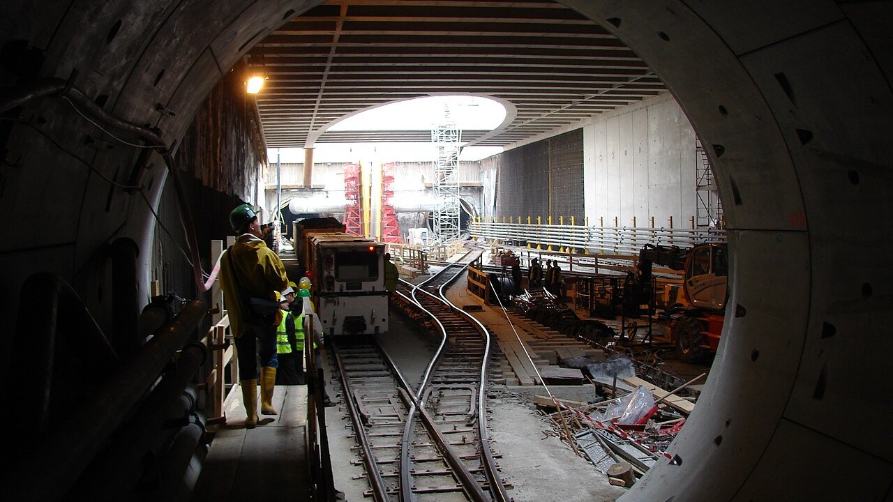 Workers and a small rail vehicle on tracks inside a concrete-lined underground metro tunnel under construction.