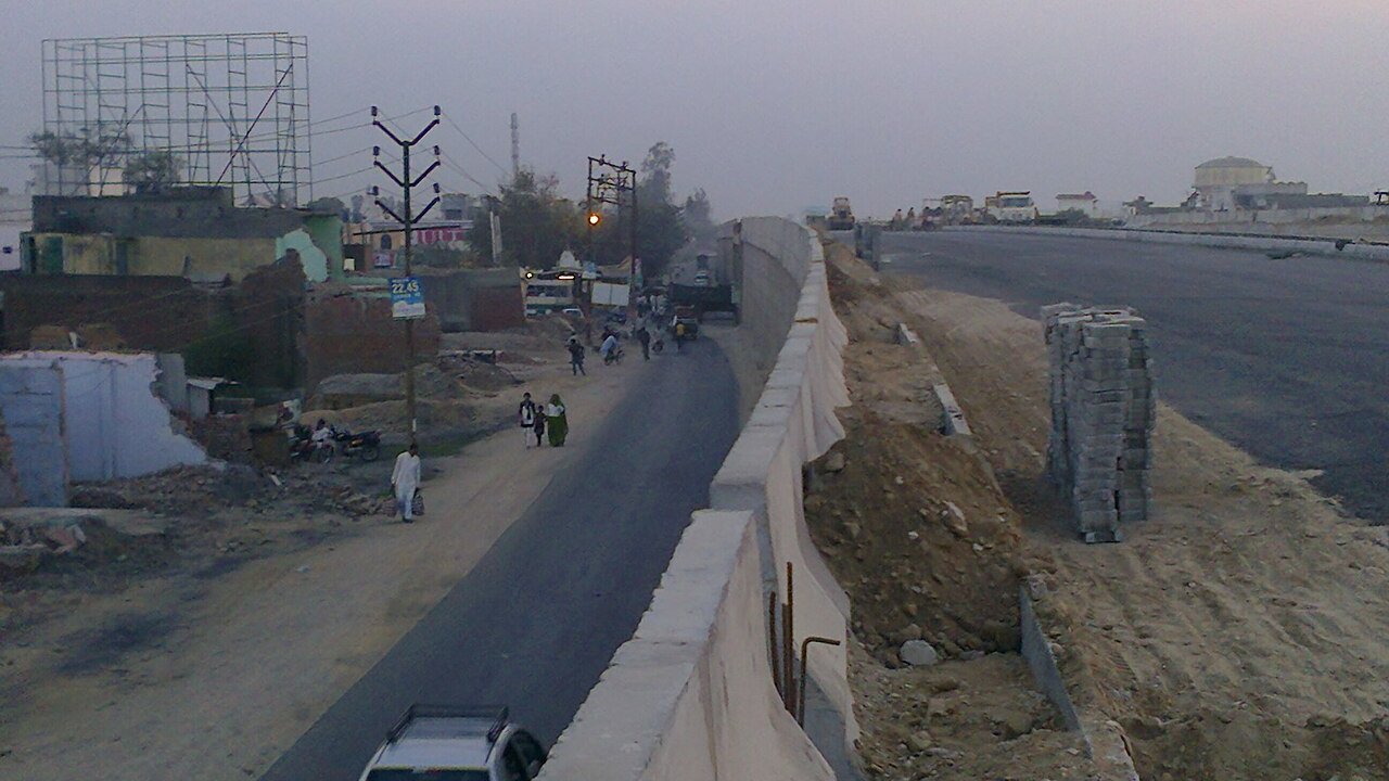 An elevated road under construction with concrete crash barriers, fresh asphalt, and people walking on the road below.