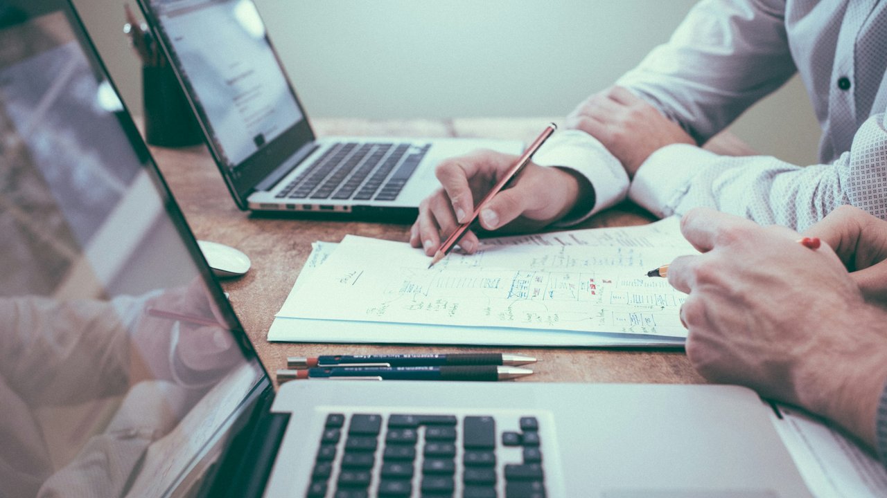 A hand holding a pencil over papers near a laptop.