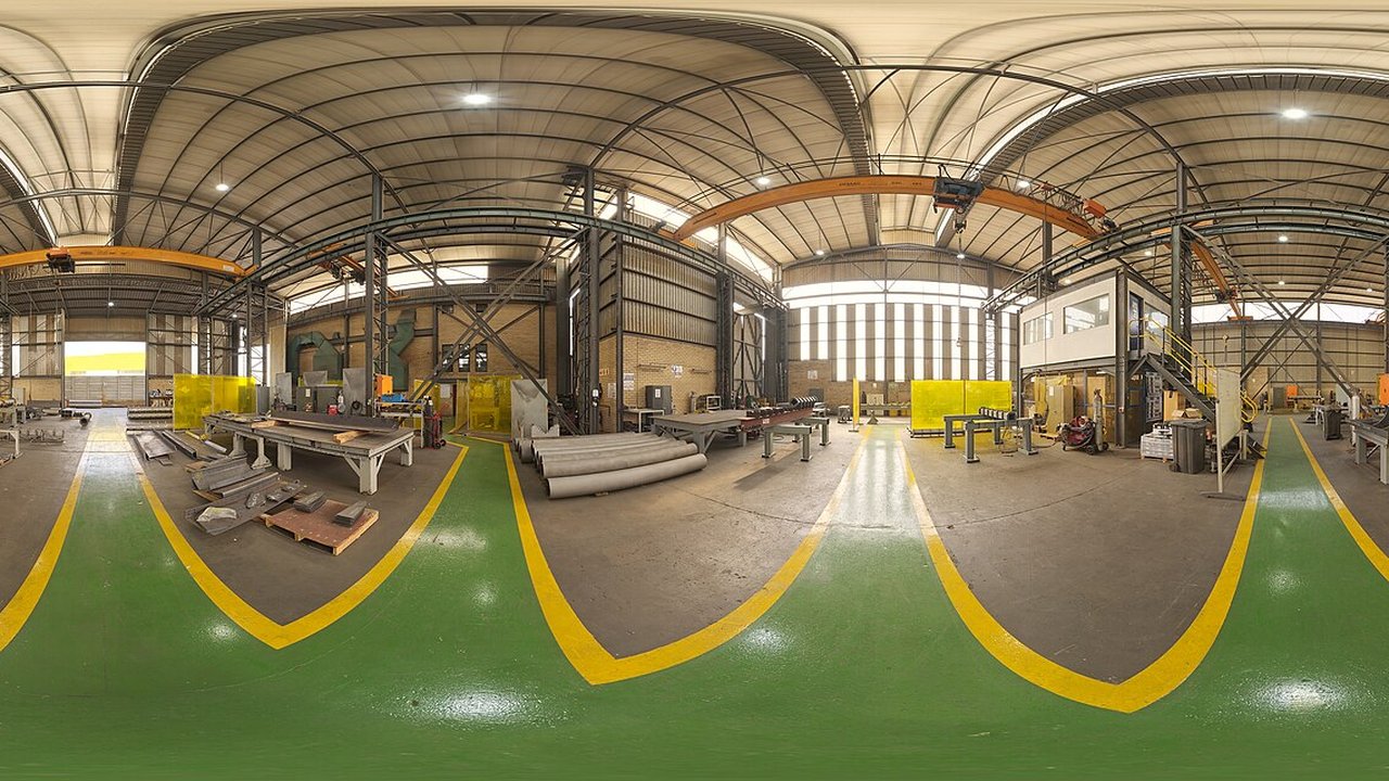 A wide industrial metal workshop with overhead cranes, steel sections stacked on workbenches, and yellow floor markings.