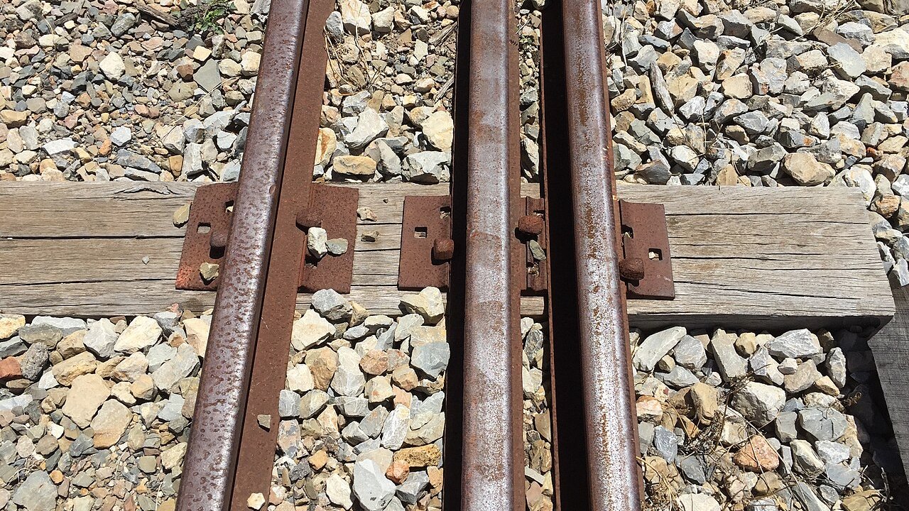 Close view of steel railway rails with rusty fittings on timber sleepers in a bed of crushed stone.