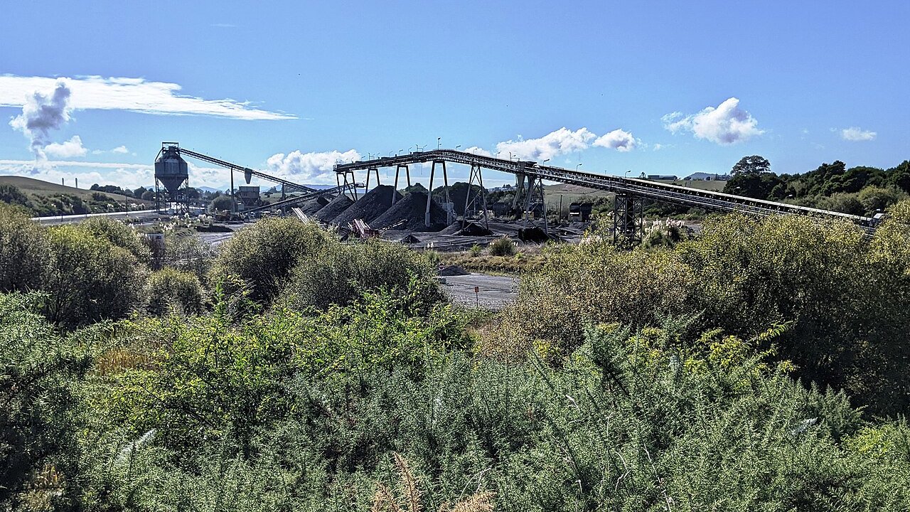 An open-cast mine site with a long overhead conveyor belt carrying material to stockpiles, surrounded by green shrubs.