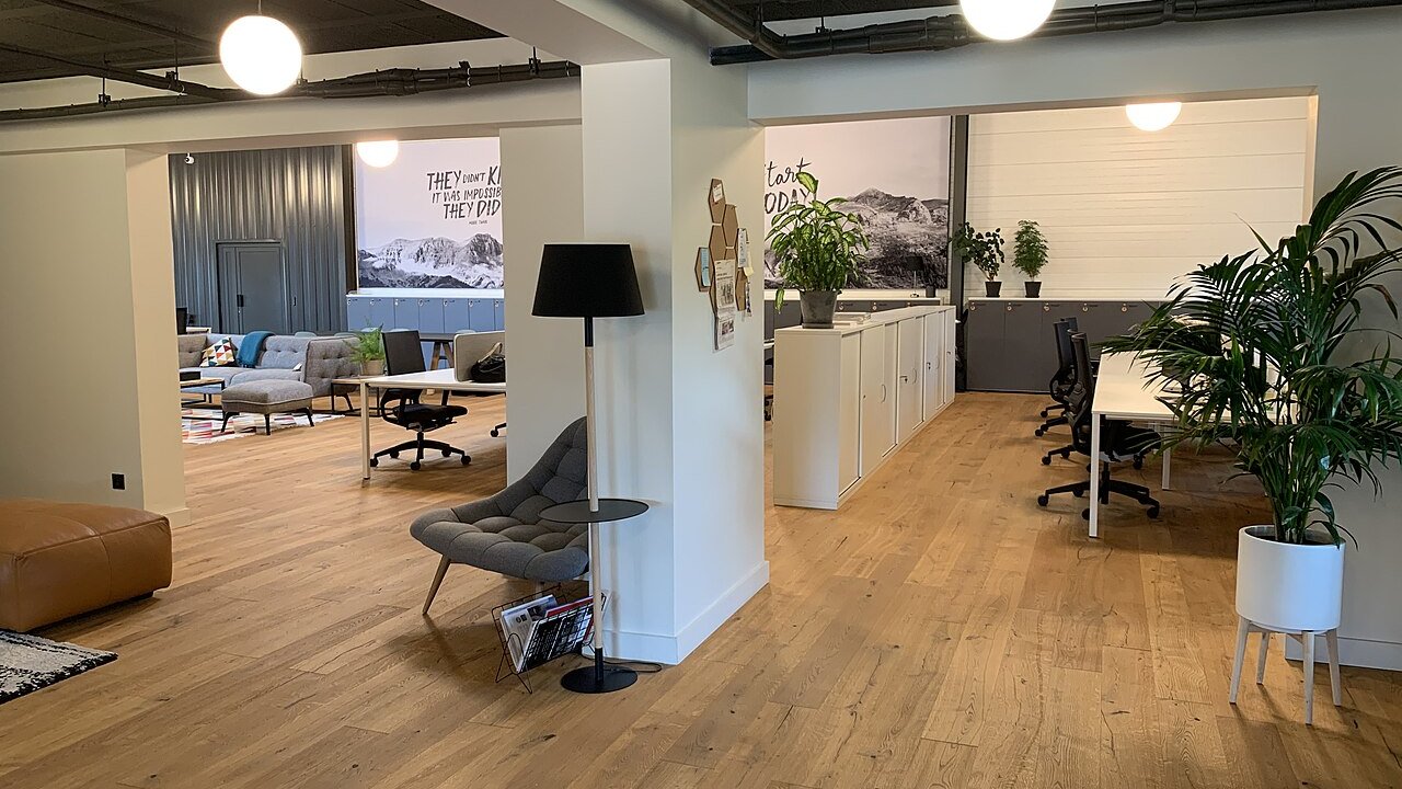 An open-plan office interior with desks, lounge seating, wall cabinets and potted plants on a wooden floor.