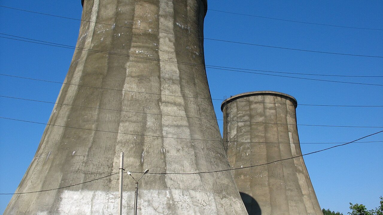 Two tall concrete cooling towers of a power station under a clear blue sky, with overhead power lines crossing in front.