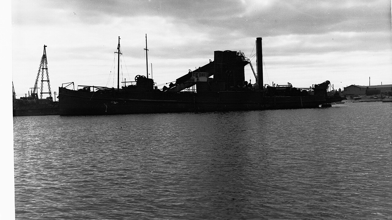 A dark black and white photograph of a dredging ship with a tall funnel and a crane-like boom moored on calm water beside a quay.