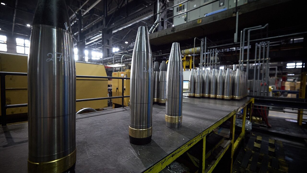 Polished steel artillery shells with brass bands stand on a metal workbench inside an industrial factory hall.