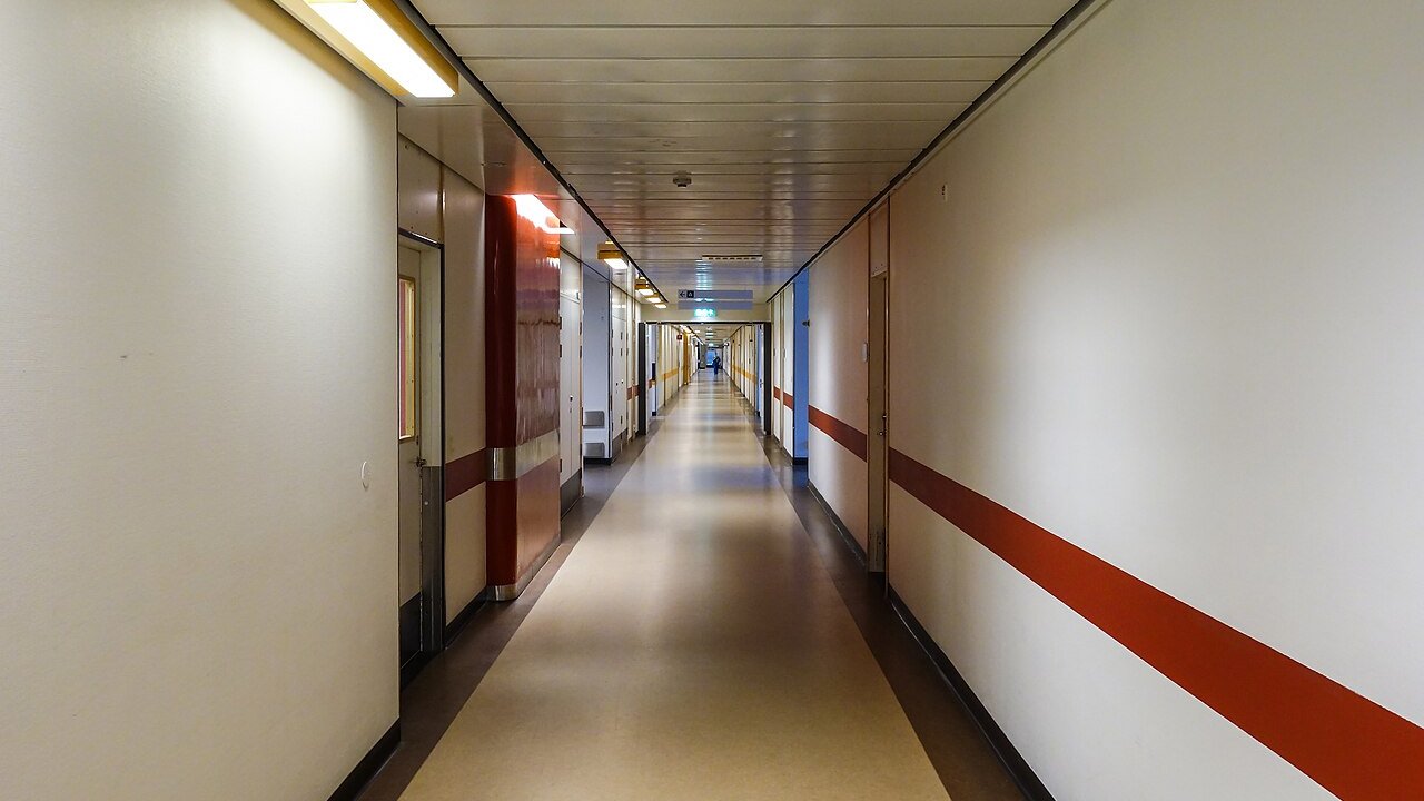 A long empty hospital corridor with cream walls, red stripes along the wall and doors on both sides.