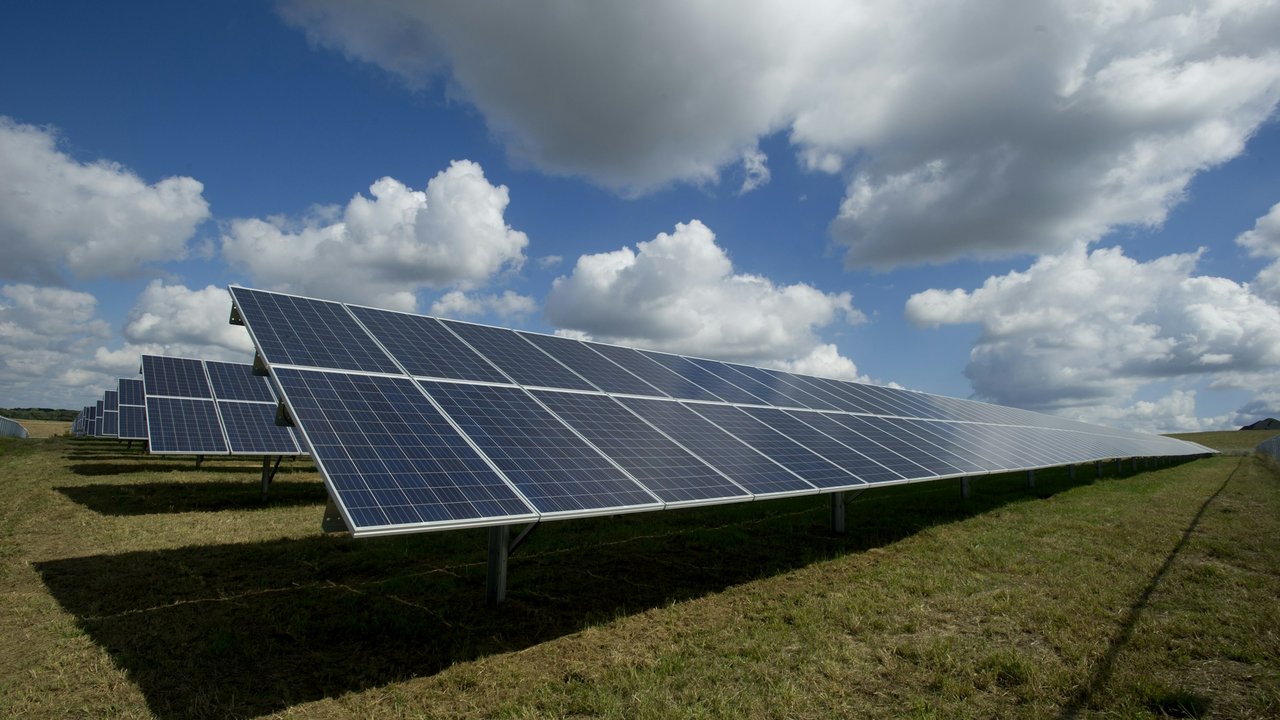 Rows of solar panels on a grassy field under a cloudy blue sky.