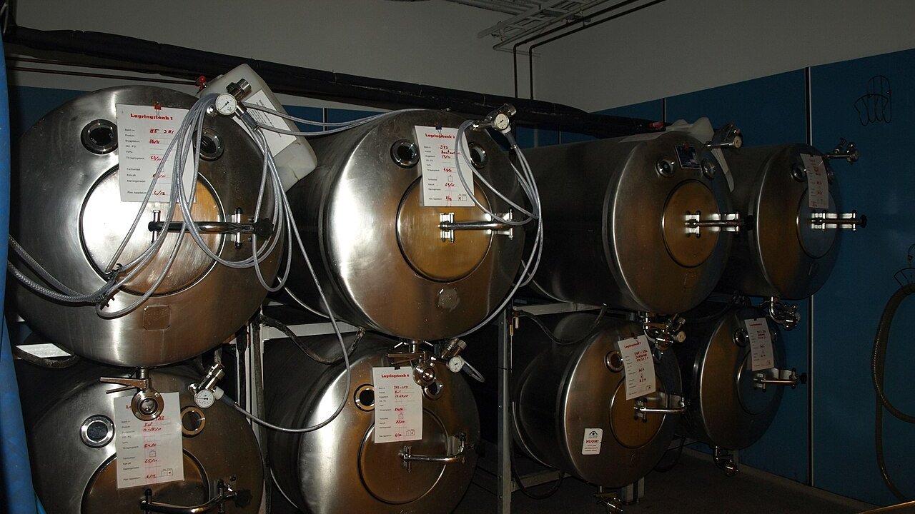 Stainless steel fermentation tanks stacked in a row, with pipes, valves and small paper tags on them.