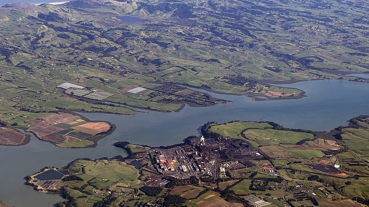 An aerial view of a coastal steel mill complex with industrial buildings and stockpiles beside green farmland and a waterway.