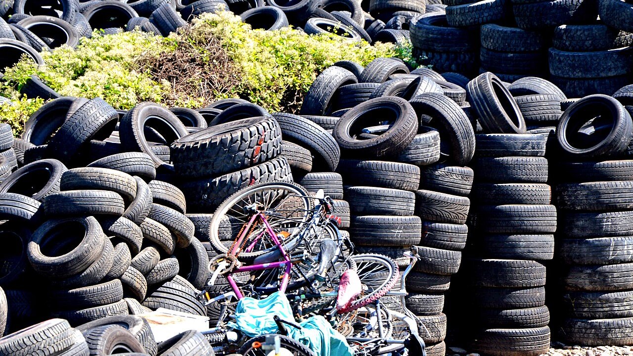 Stacks of used tyres and a pile of abandoned bicycles at a recycling yard, with green shrubs behind.