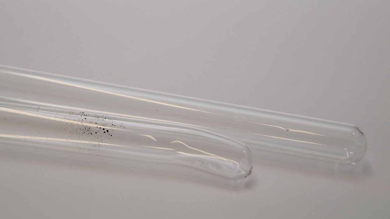 Two empty glass test tubes lying side by side on a plain grey surface, with a few dark specks in one tube.