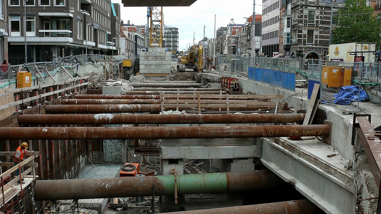 A deep urban excavation with steel pipes braced across a construction pit, a worker in a high-visibility vest, and an excavator and crane in the background.