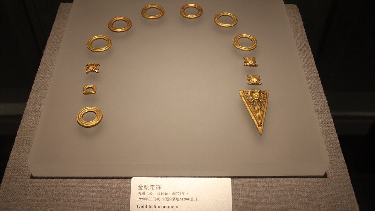 Ornate gold belt ornaments, rings and fittings, displayed on a grey pedestal with a museum caption card