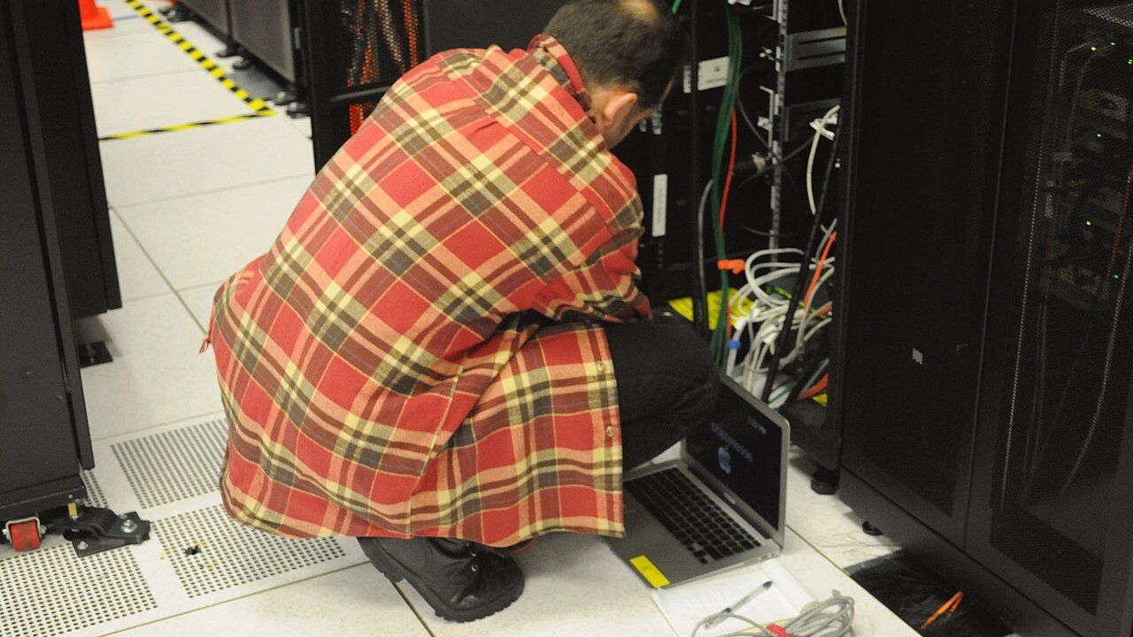 A technician in a plaid jacket crouches with a laptop beside server racks and network cabling in a data centre