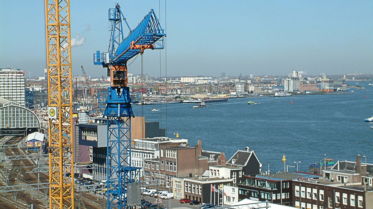 Blue and yellow construction cranes over a waterfront city with buildings and a river.