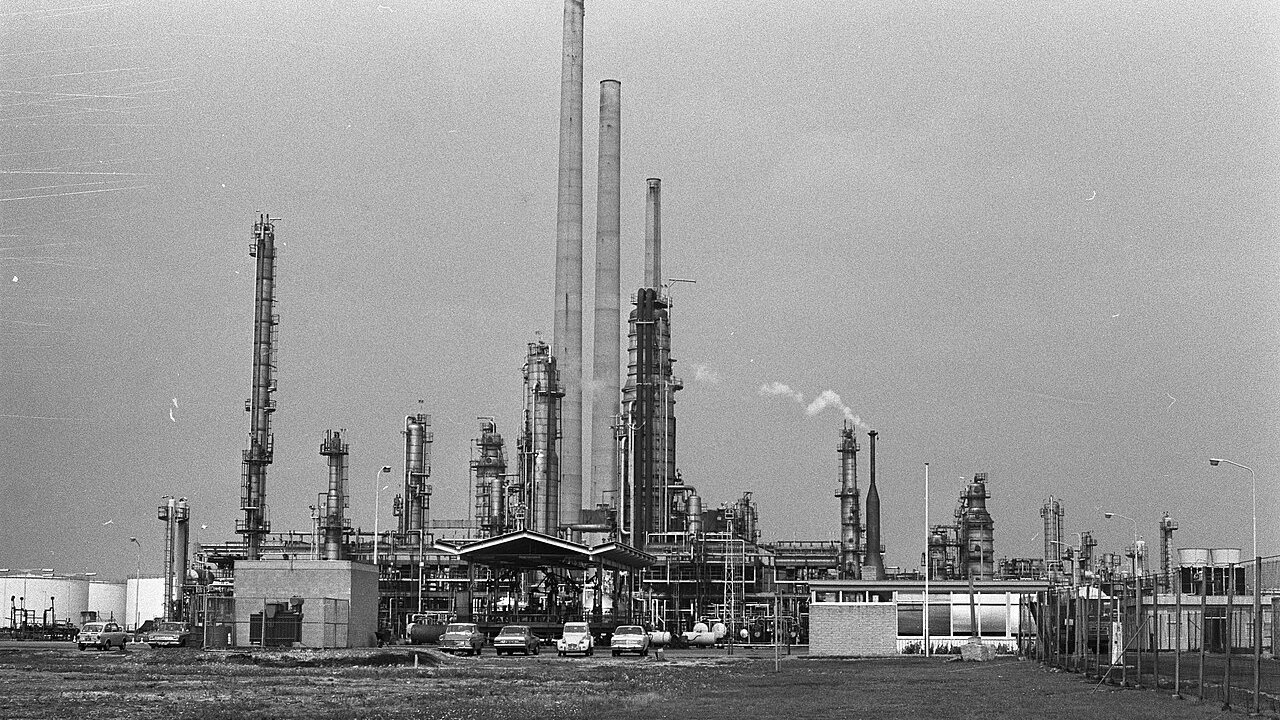 A refinery complex with tall distillation columns, chimneys and pipework in a flat, open landscape, with parked cars in the foreground.