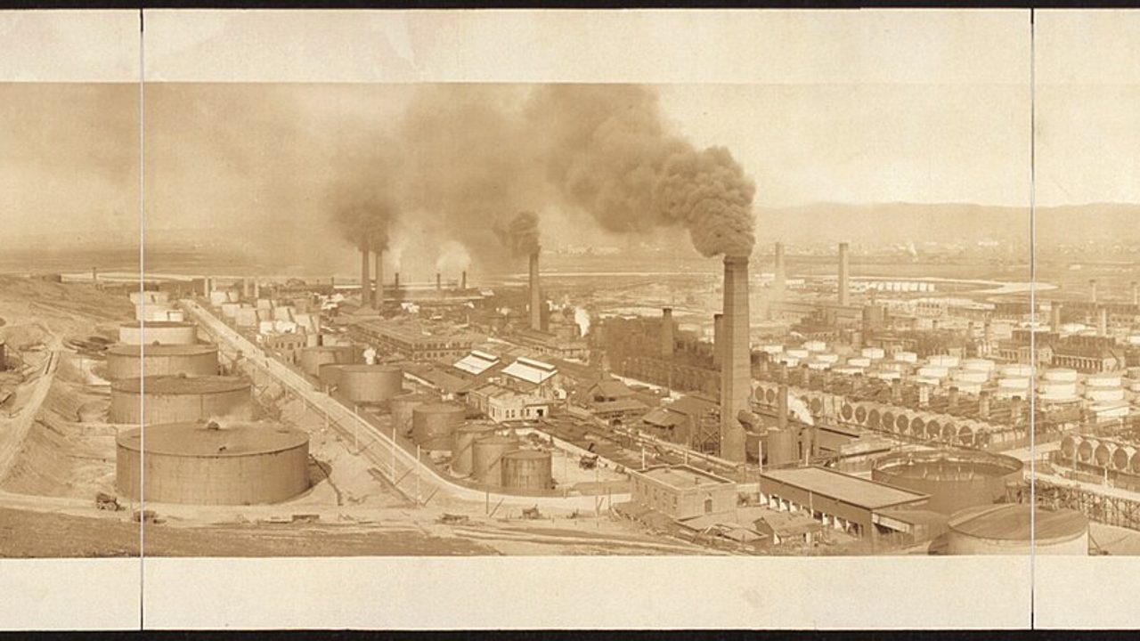 An old panoramic view of an oil refinery with storage tanks and smoking stacks