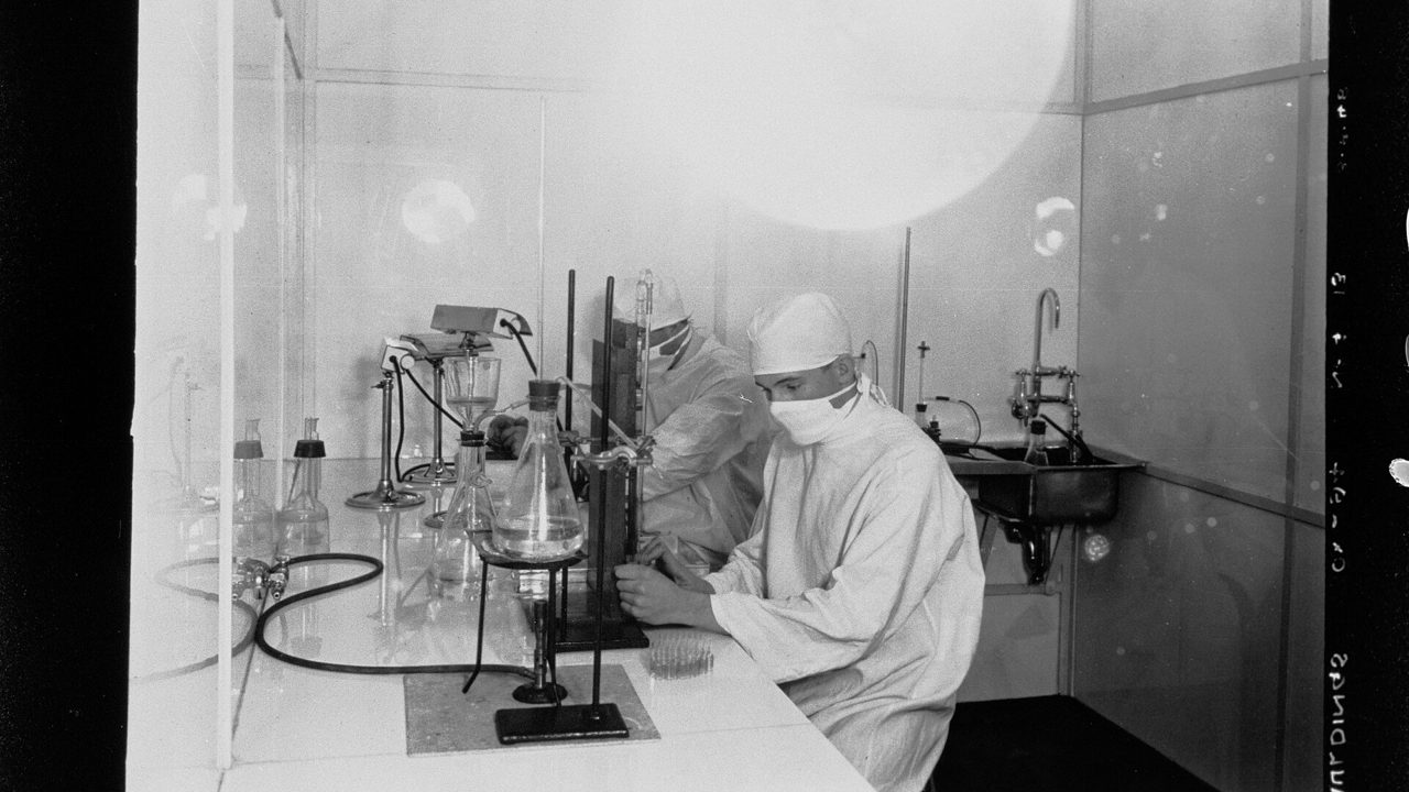 A technician in a white coat working at laboratory glassware, in an old black-and-white photograph