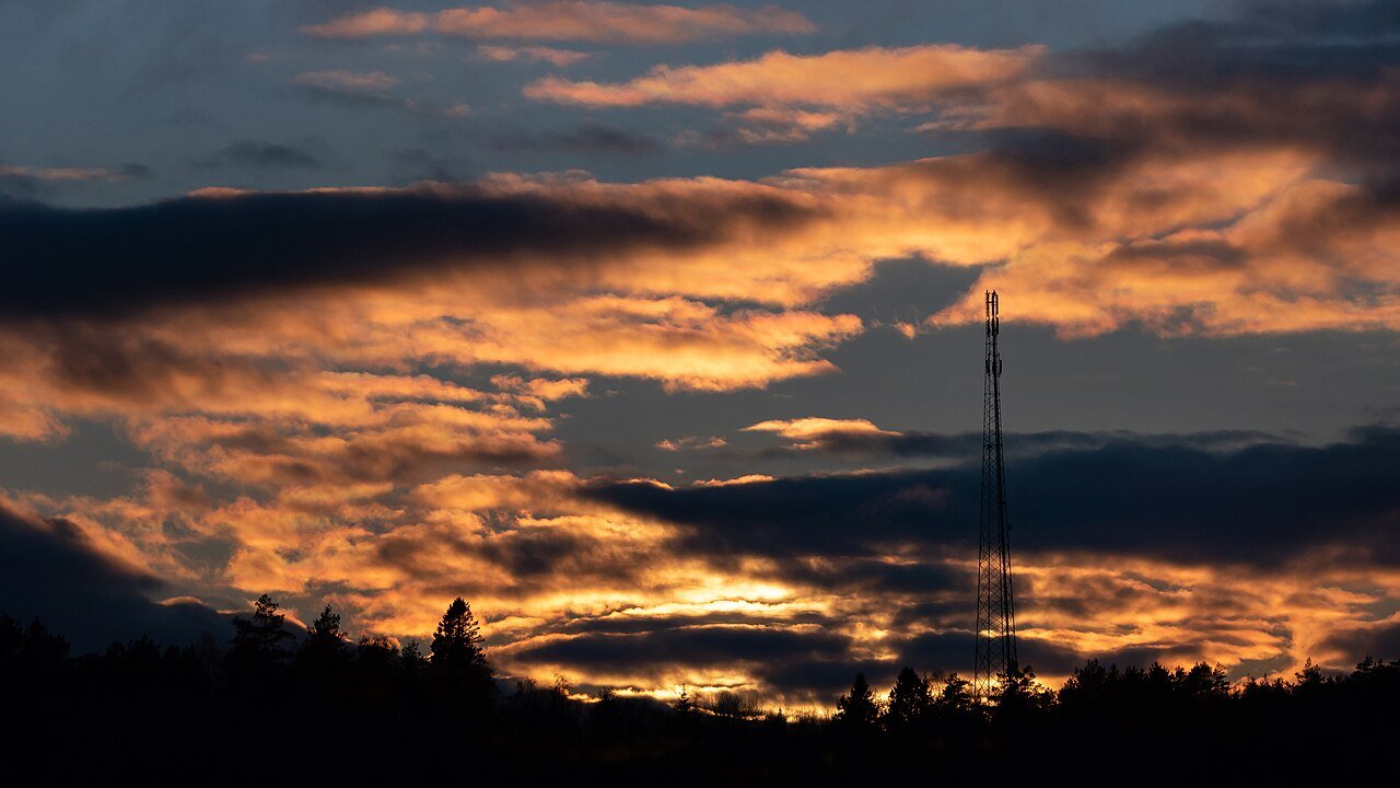 A telecom mast silhouetted against a sunset sky with orange and dark clouds above a line of trees
