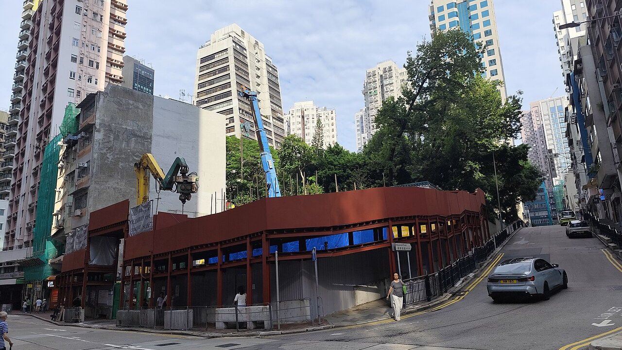 A construction site with a red steelframed temporary structure, a crane and high-rise buildings on a city street