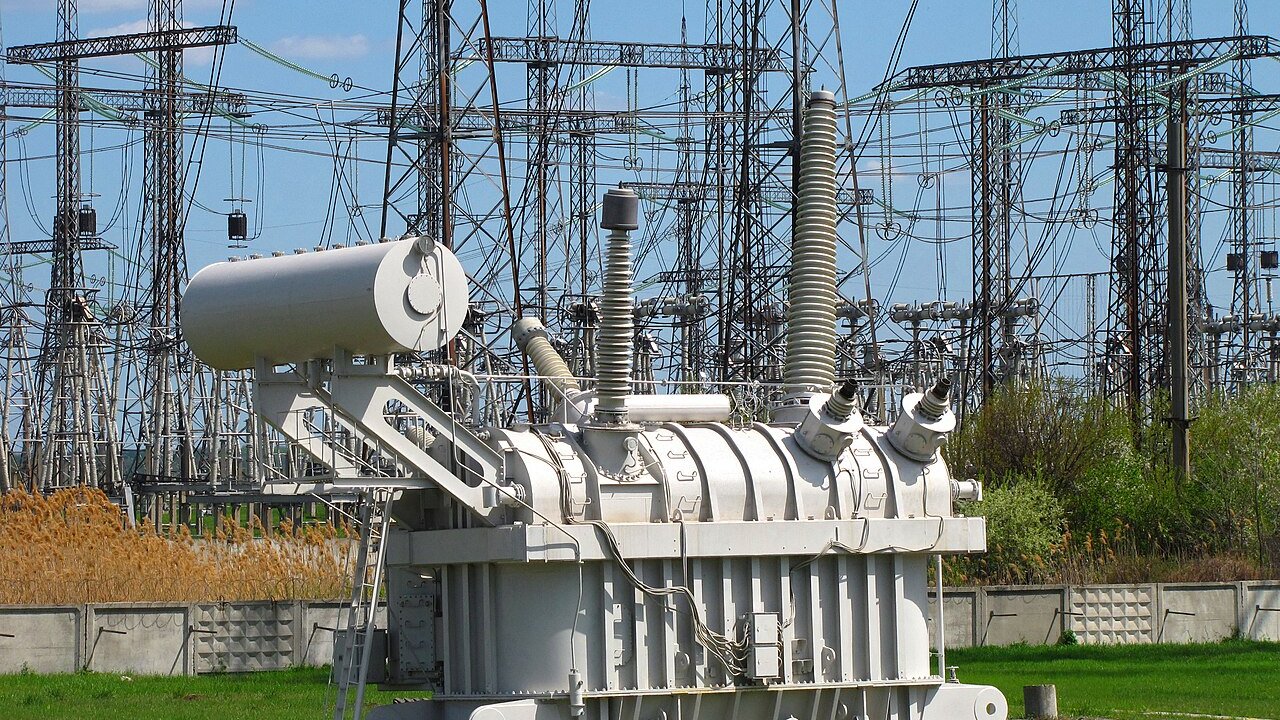 A large grey high-voltage transformer at an electrical substation with steel lattice towers and power lines behind it.