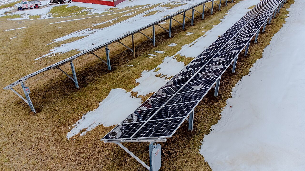 A row of ground-mounted solar panels on snowy grass beside a red building