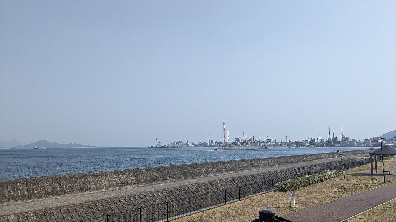 A coastal industrial complex with chimneys and plant structures across a bay, seen from a waterfront promenade