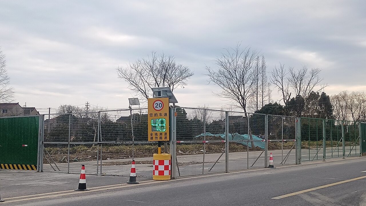 A roadside construction site behind green and wire mesh fencing, with orange and white traffic cones and a speed-limit sign with a solar-powered display.