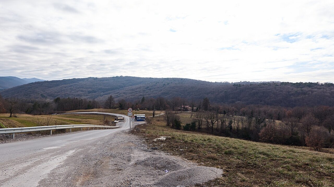 A curving unfinished road and construction site with gravel and a parked blue vehicle, set against wooded hills under an overcast sky