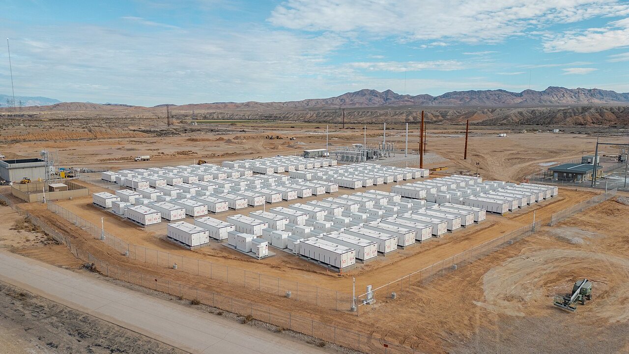 An aerial view of rows of white battery storage containers inside a fenced desert site.