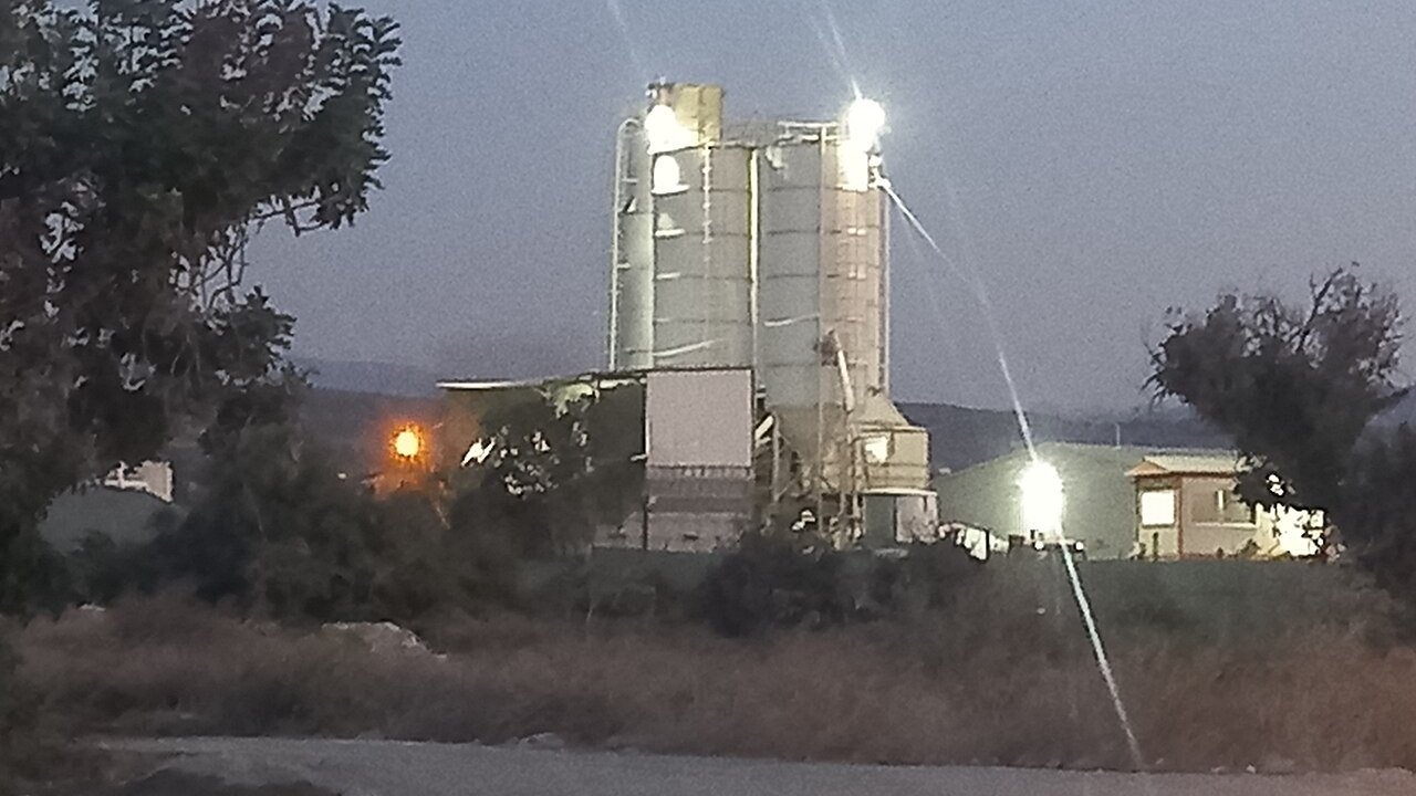 A cement plant with grey silos and a processing building lit by floodlights at dusk, with bushes in the foreground.