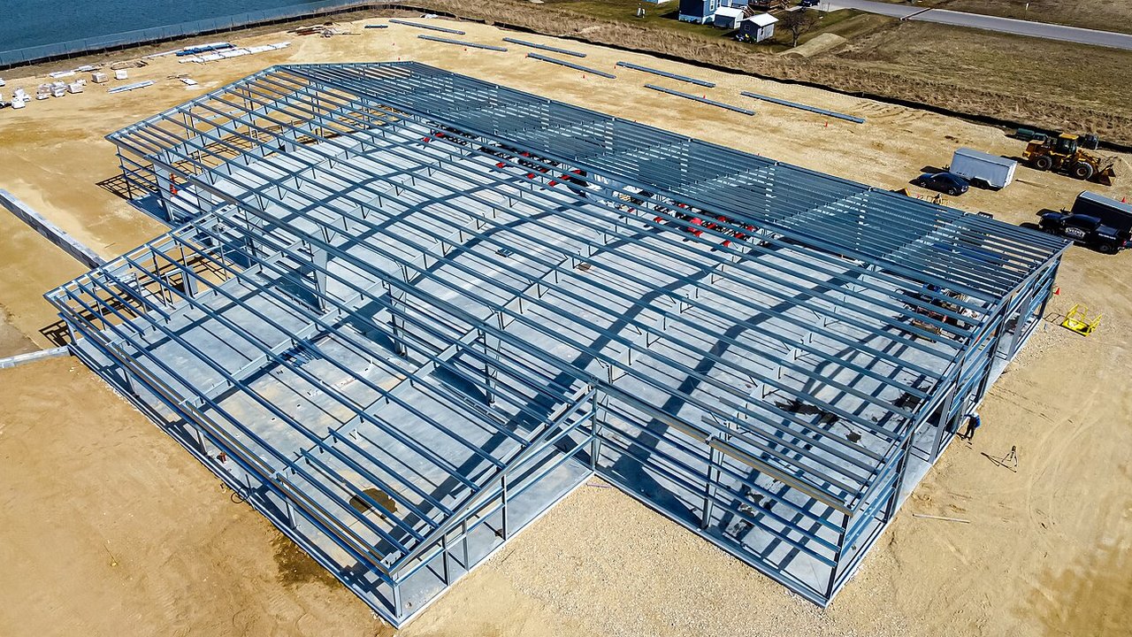 A large steel-frame building under construction on a cleared site, with its galvanised roof and wall framing exposed.