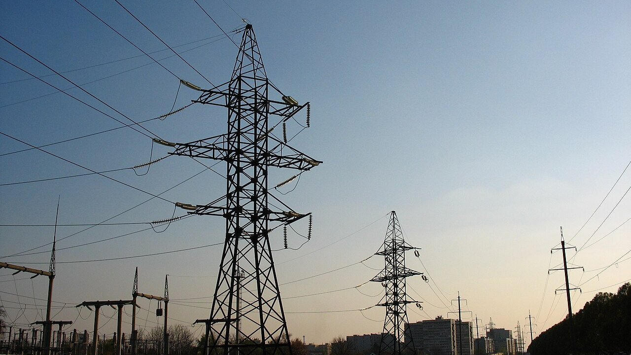 High-voltage steel transmission towers and overhead power lines against a clear evening sky, with substation equipment and city buildings in the distance