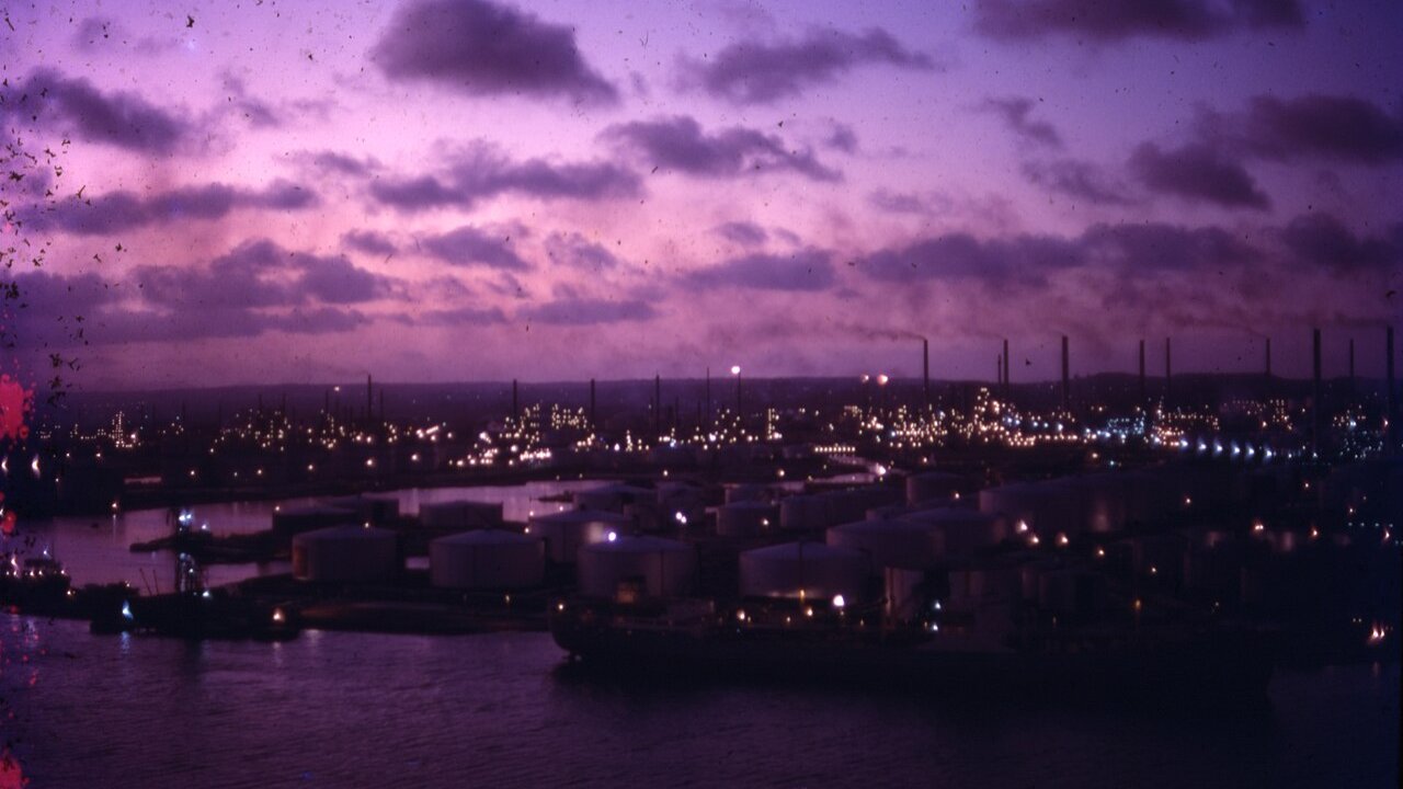A dusk view of an oil refinery with rows of storage tanks beside a waterway and lit industrial plant beyond