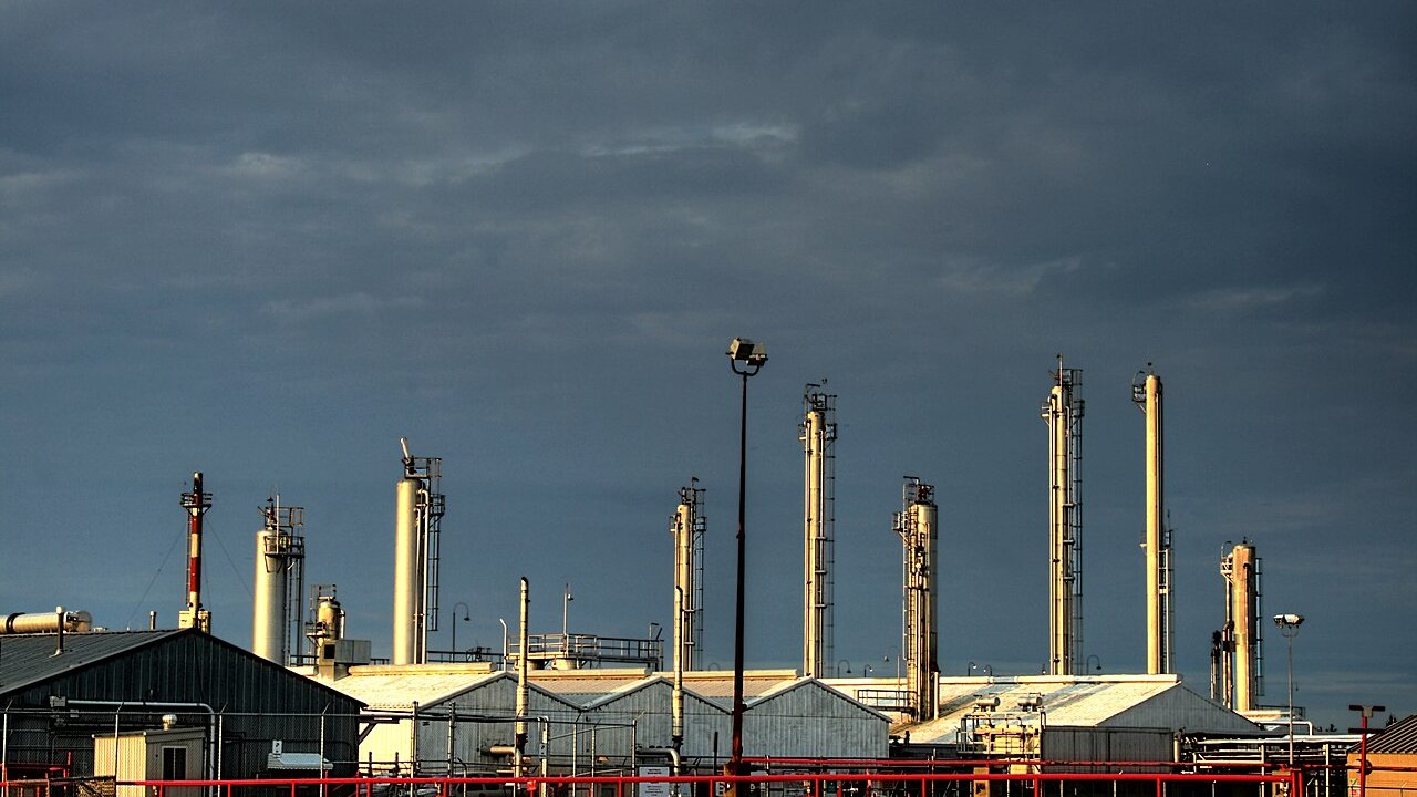 A petrochemical plant with tall process stacks, pipework and industrial sheds under a dark cloudy sky