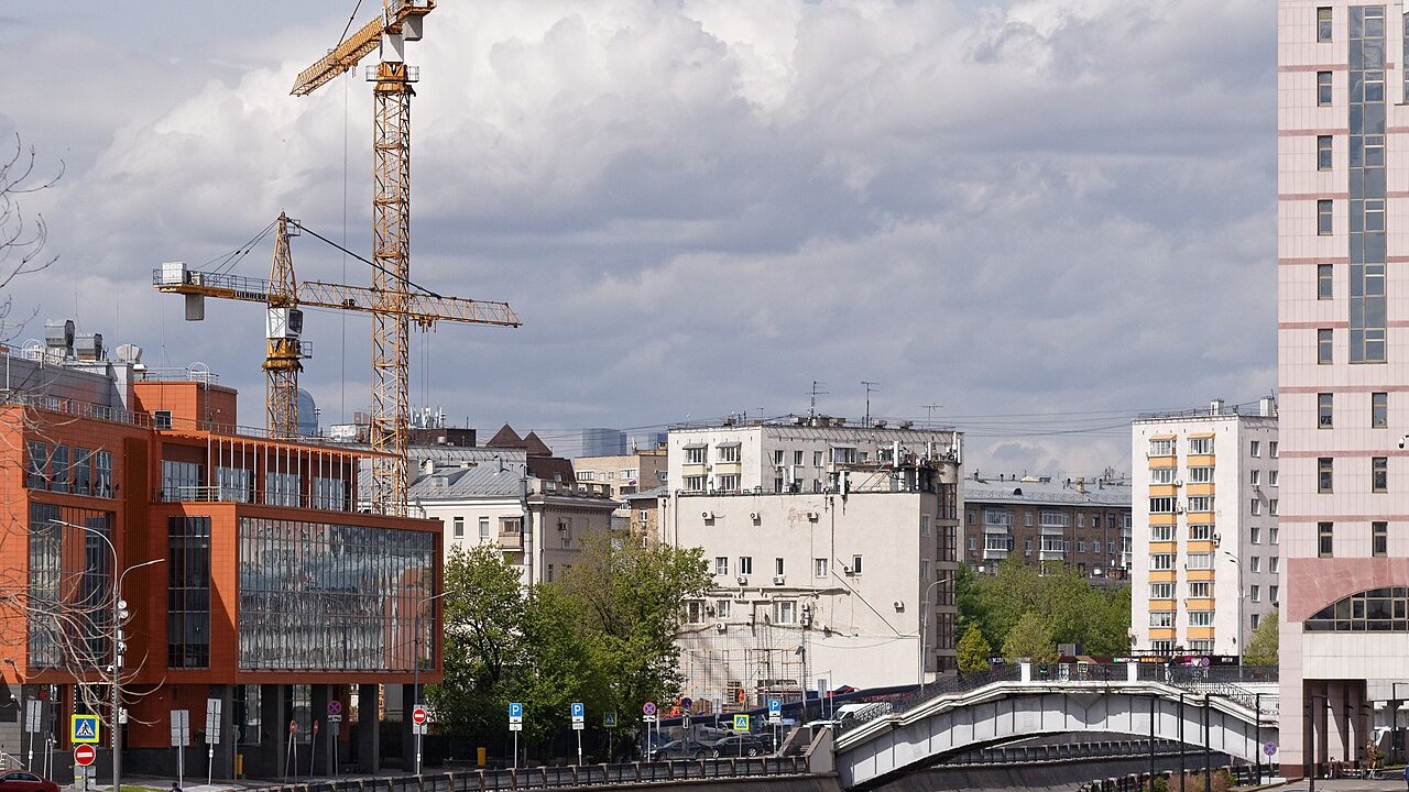 Tower cranes over a city street with mid-rise apartment and office buildings and a footbridge in the foreground